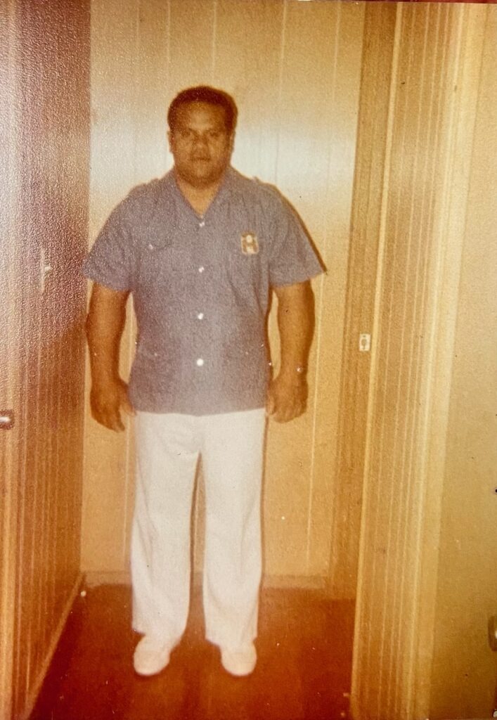 A man stands in a doorway wearing a blue shirt and white pants, exuding confidence in the home setting. The photo captures a vintage style.