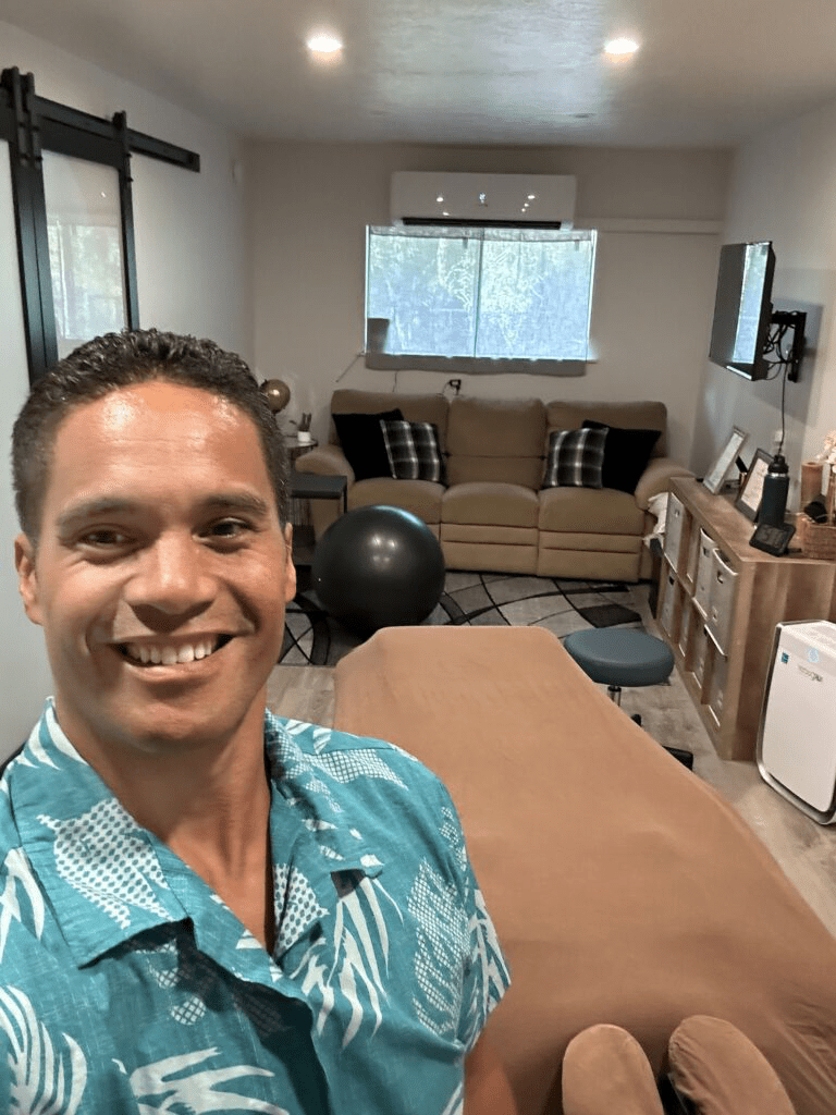 A person smiling in a turquoise shirt stands in a cozy therapy room featuring a massage table, couch, and exercise ball, indicating a wellness environment.