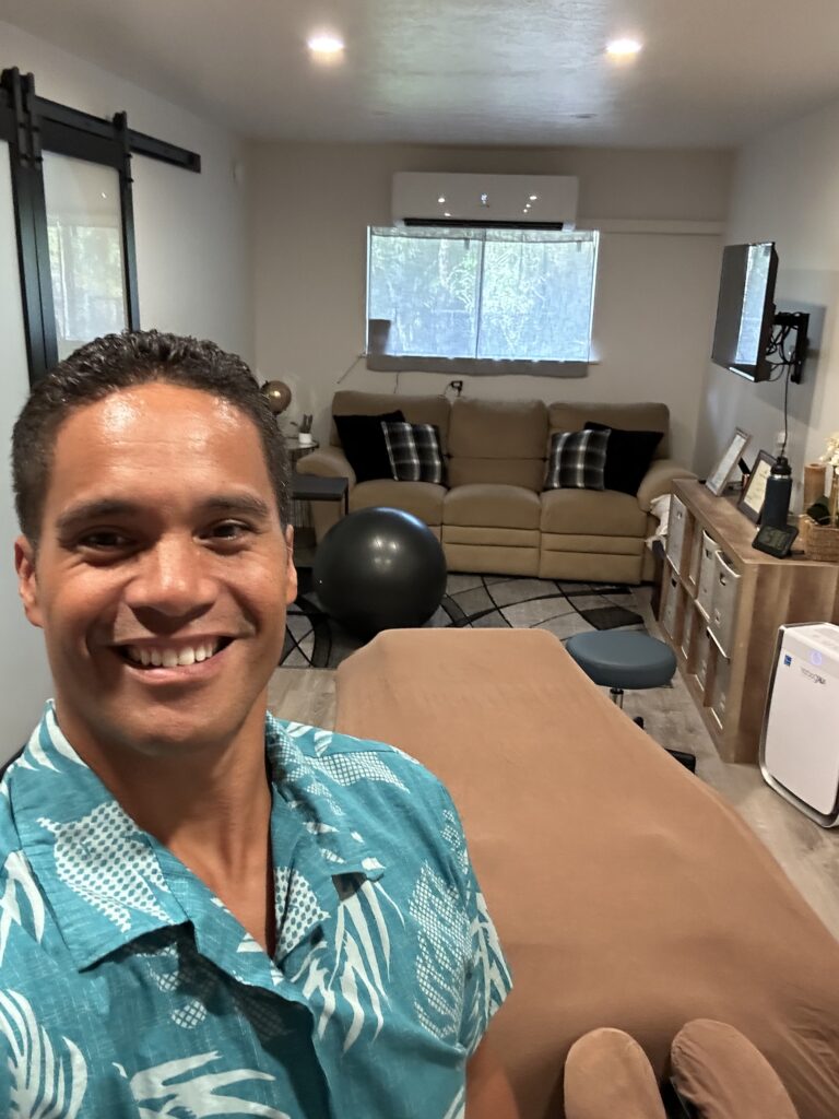 Wellness practitioner smiling in a therapy room with a massage table, sofa, and exercise ball, highlighting a cozy, inviting atmosphere.