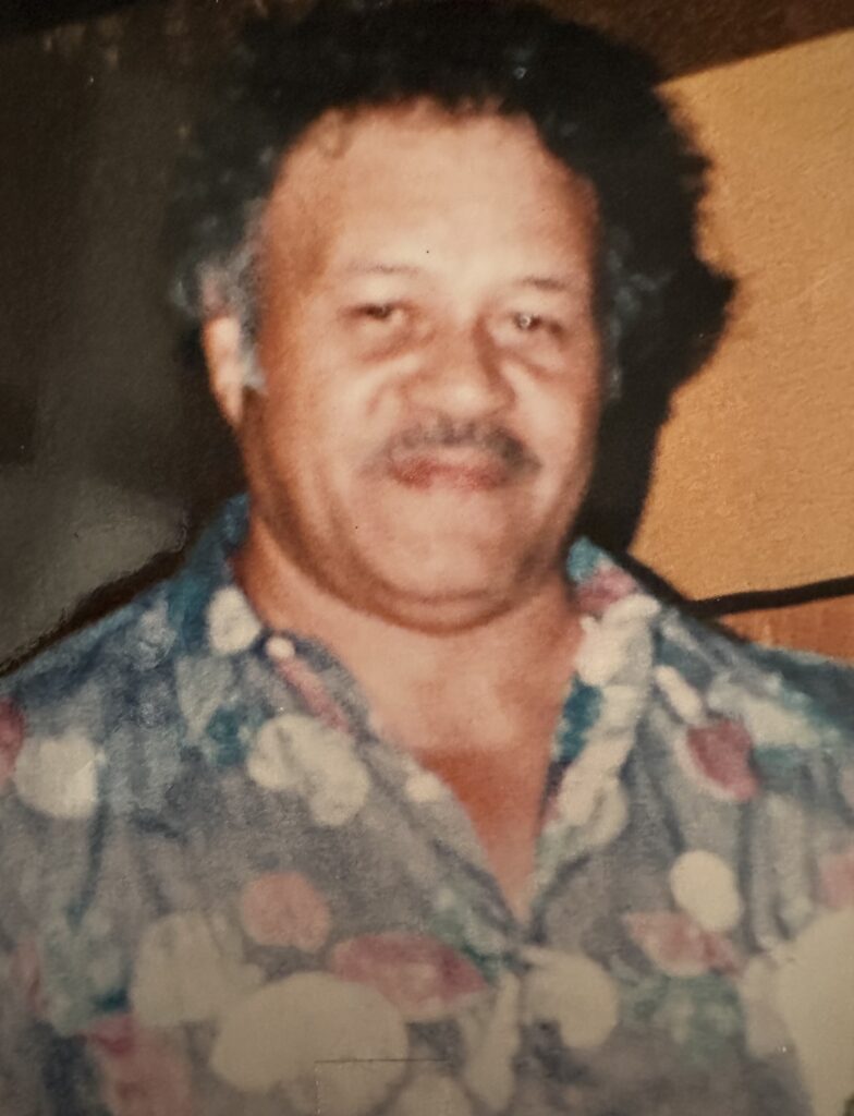 A middle-aged man with curly black hair smiles, wearing a colorful floral shirt against a muted background, exuding warmth and friendliness.