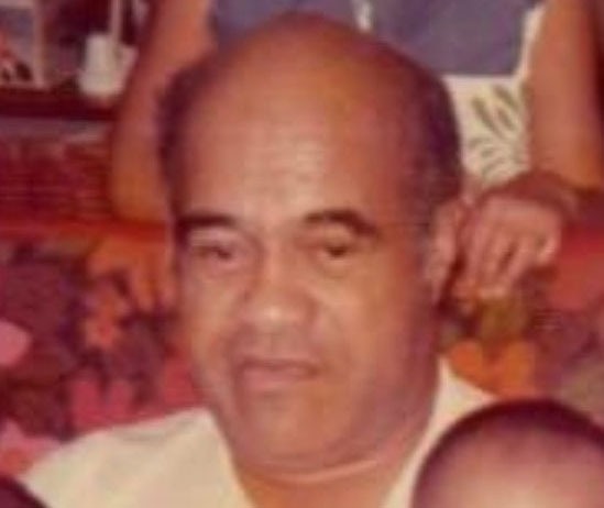 A middle-aged man with a bald head and serious expression sits in front of a vibrant floral backdrop, suggesting a familial gathering or celebration.