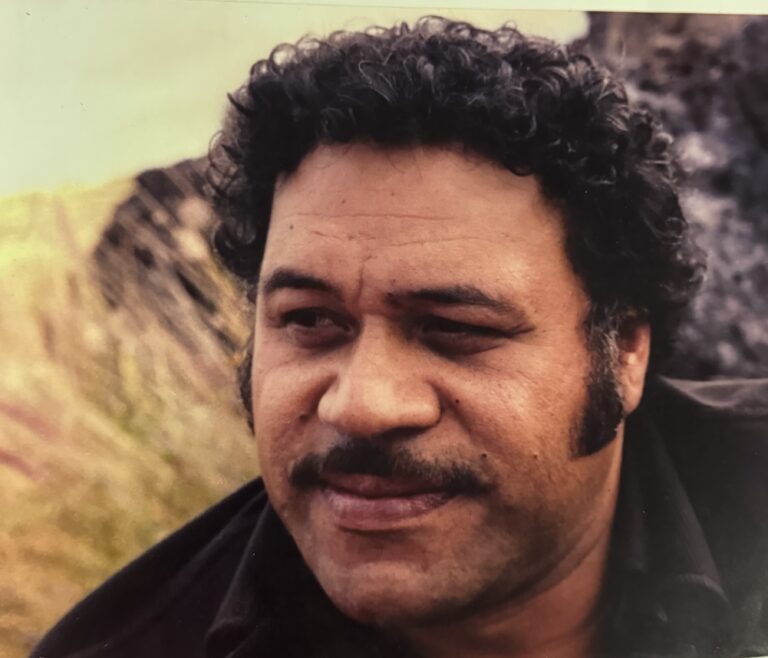Man with curly hair and a mustache smiles thoughtfully outdoors, surrounded by a natural landscape. The image evokes warmth and contemplation.