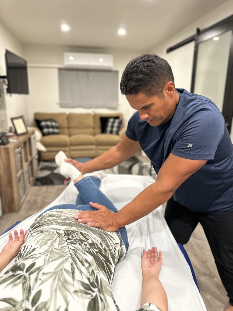 A male therapist assists a patient on a treatment table, providing care on her lower back in a cozy, neutral-toned room.